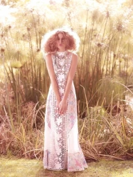 Model with voluminous curly red hair wearing a sleeveless patterned white maxi dress standing outdoors among tall reeds.