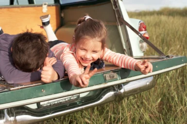 Two children lying in the open tailgate of a vintage station wagon in a grassy field