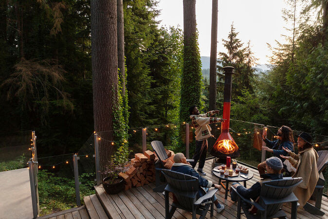 Group of people on a wooden deck near a modern outdoor fireplace in a forest setting, enjoying drinks and conversation.