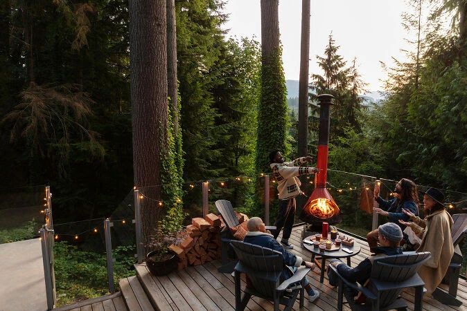 Group of people on a wooden deck near a modern outdoor fireplace in a forest setting, enjoying drinks and conversation.