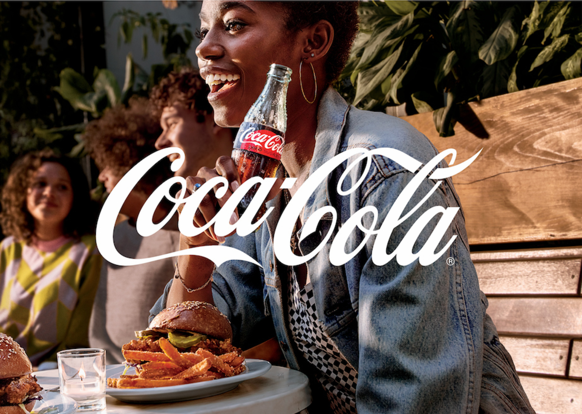 A cheerful African-American woman smiles while drinking Coca-Cola in a sunny outdoor setting with friends and delicious-looking food.
