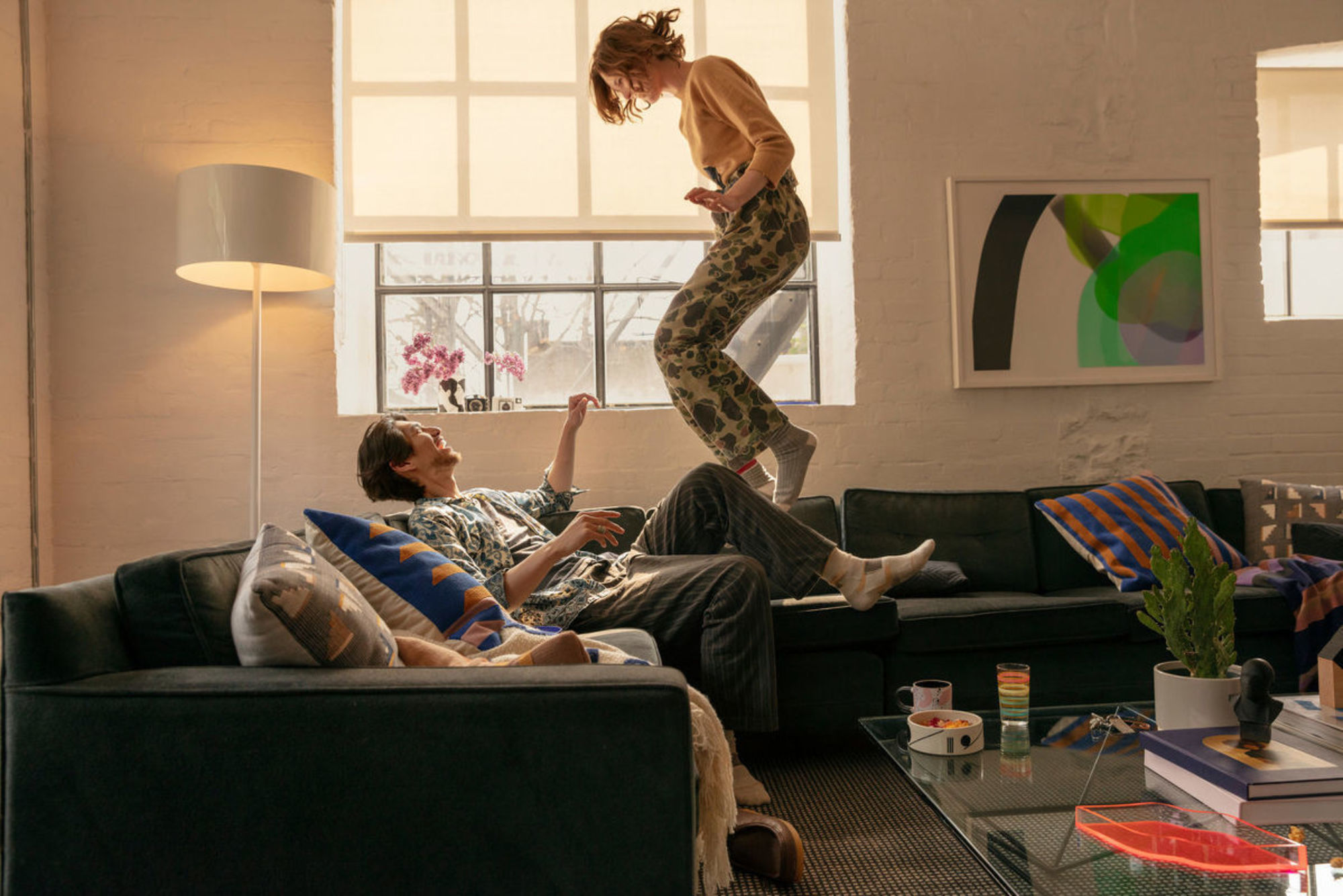 A woman stands on a window ledge behind a man reclining on a couch in a brightly lit room. Colorful pillows and modern art.