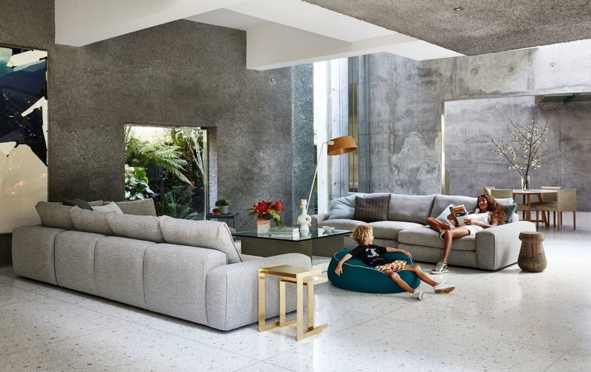 Contemporary living room interior with gray concrete walls, modern furniture, a woman reading on a sofa, and a boy on a beanbag.