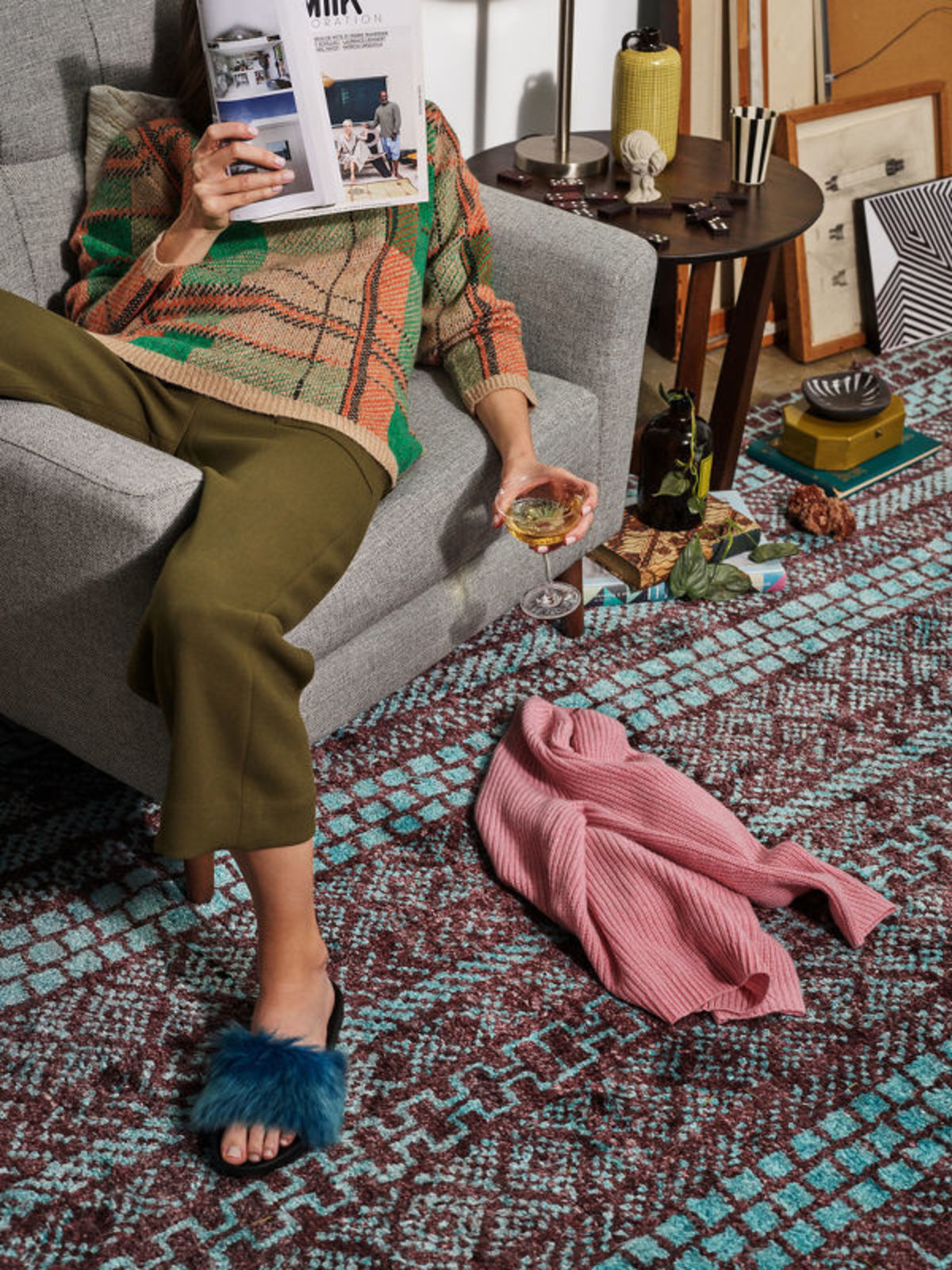 A woman relaxes in a gray armchair, reading a magazine and sipping wine, with a colorful rug and stylish decor around her.