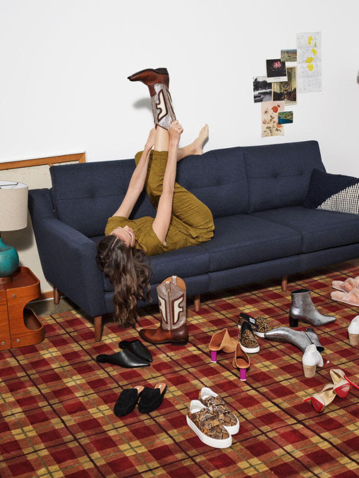 Adult woman lying on a blue couch, wearing a yellow outfit and one brown cowboy boot, surrounded by an assortment of shoes on a plaid floor.
