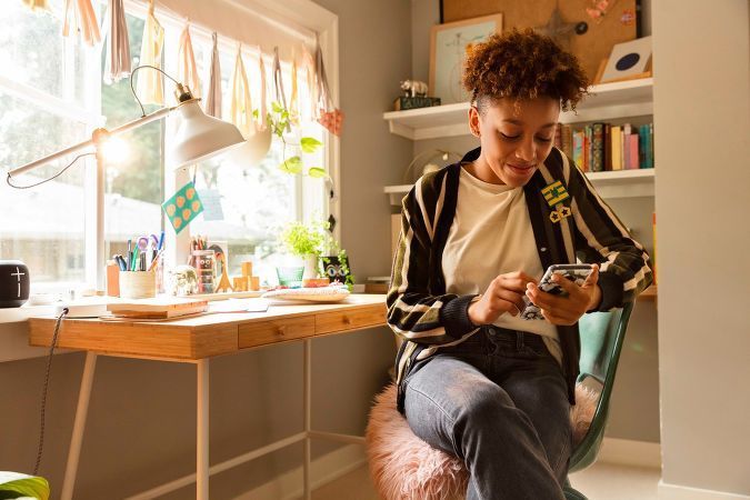 Woman seated at desk in bright room using smartphone. Natural light fills the space with books, art, and decor accents.