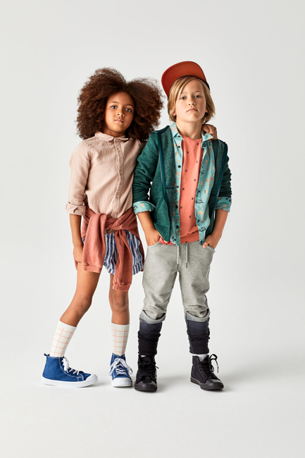 Two children, a girl with afro hair and a boy wearing a cap, stand side by side in a studio shot. Both are dressed in casual stylish clothing.