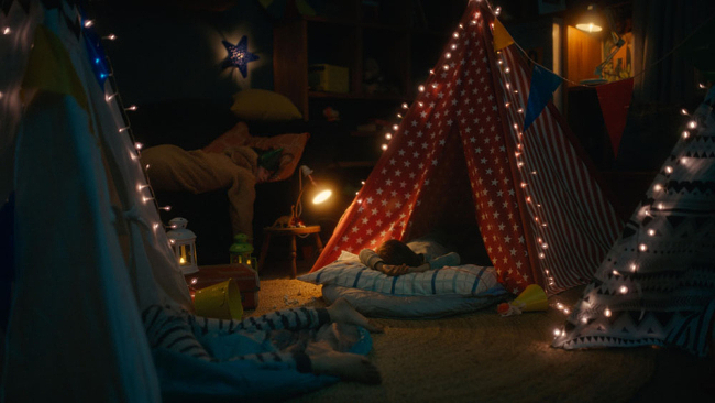 Kids create a cozy indoor campsite with homemade tents, string lights, pillows and blankets for a fun family adventure at home.