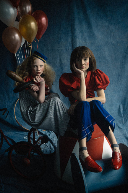 Two young models pose indoors on a striped platform, surrounded by vintage bicycles and balloons, wearing red and blue outfits against a blue backdrop.
