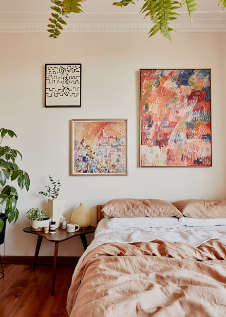 Bedroom interior with a made bed, abstract paintings on the wall, a small table with plants and decorative objects, and hanging ferns.