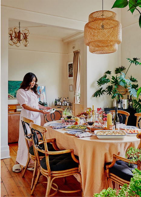 Woman in pink loungewear sets white dishes on a dining table with food and wine, surrounded by plants in a sunlit room.