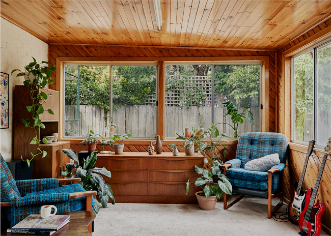 Living room interior with wood paneled walls, blue plaid armchairs, plants, and natural light from a window overlooking a garden.