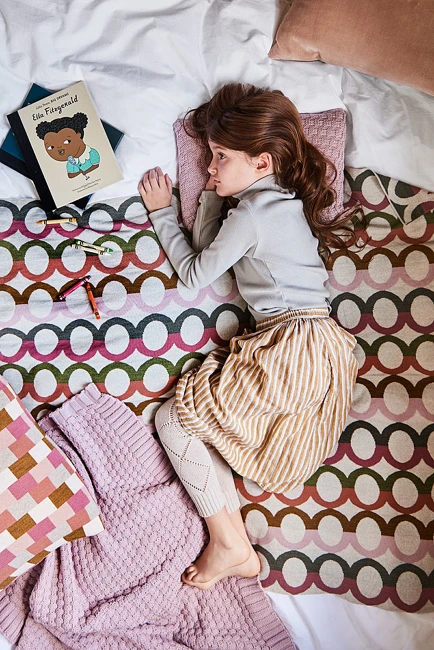 A young child lying on a patterned blanket with a book, pillows, and scattered crayons on a bed.