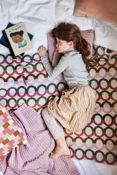 A young child lying on a patterned blanket with a book, pillows, and scattered crayons on a bed.