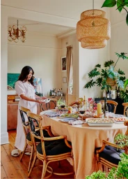 A woman in a pink dress standing beside a long dining table set with food and wine inside a bright living space