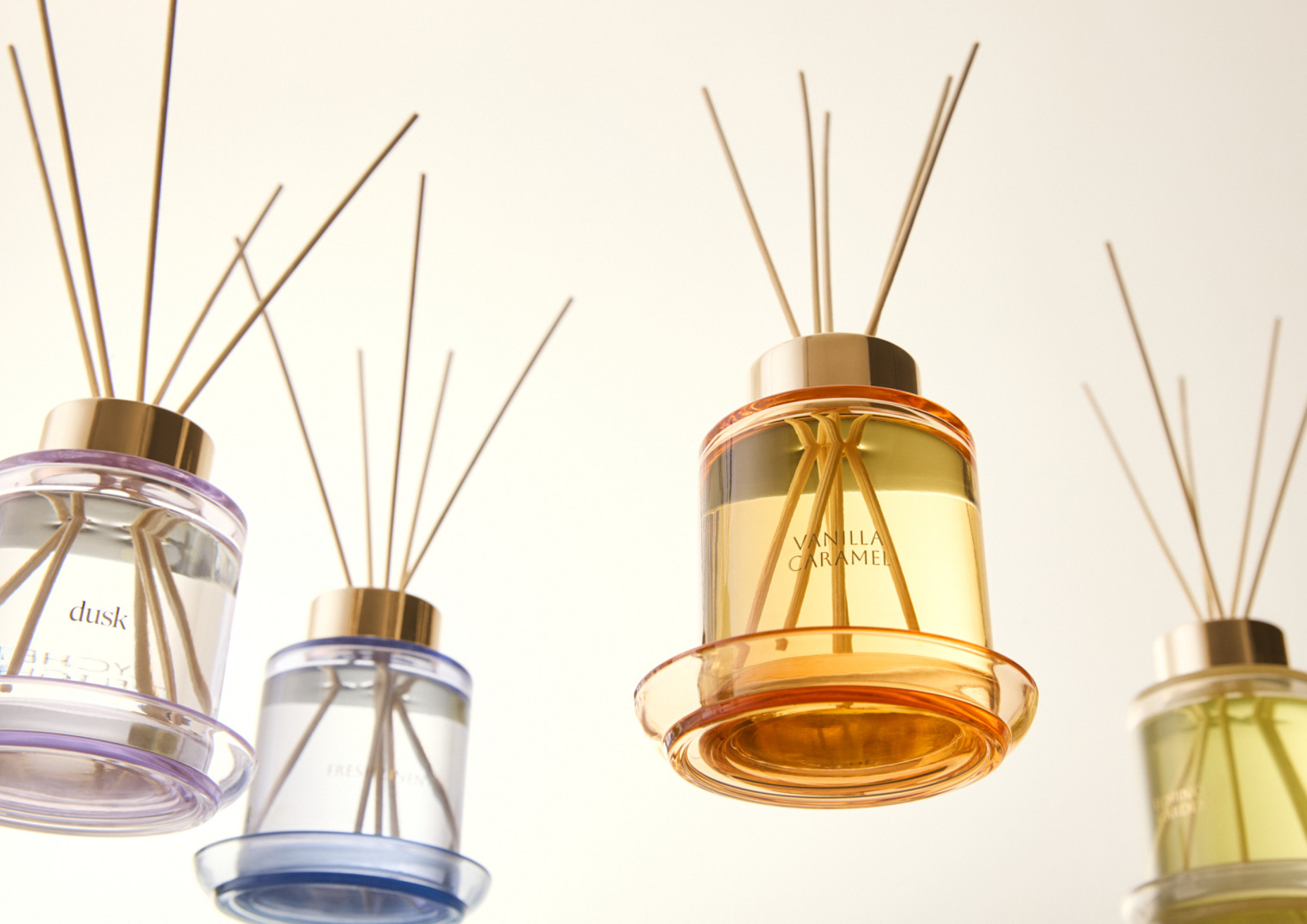 Five glass reed diffusers in varying colors, from left to right: purple, blue, orange, and green, with wooden reeds sticking out.