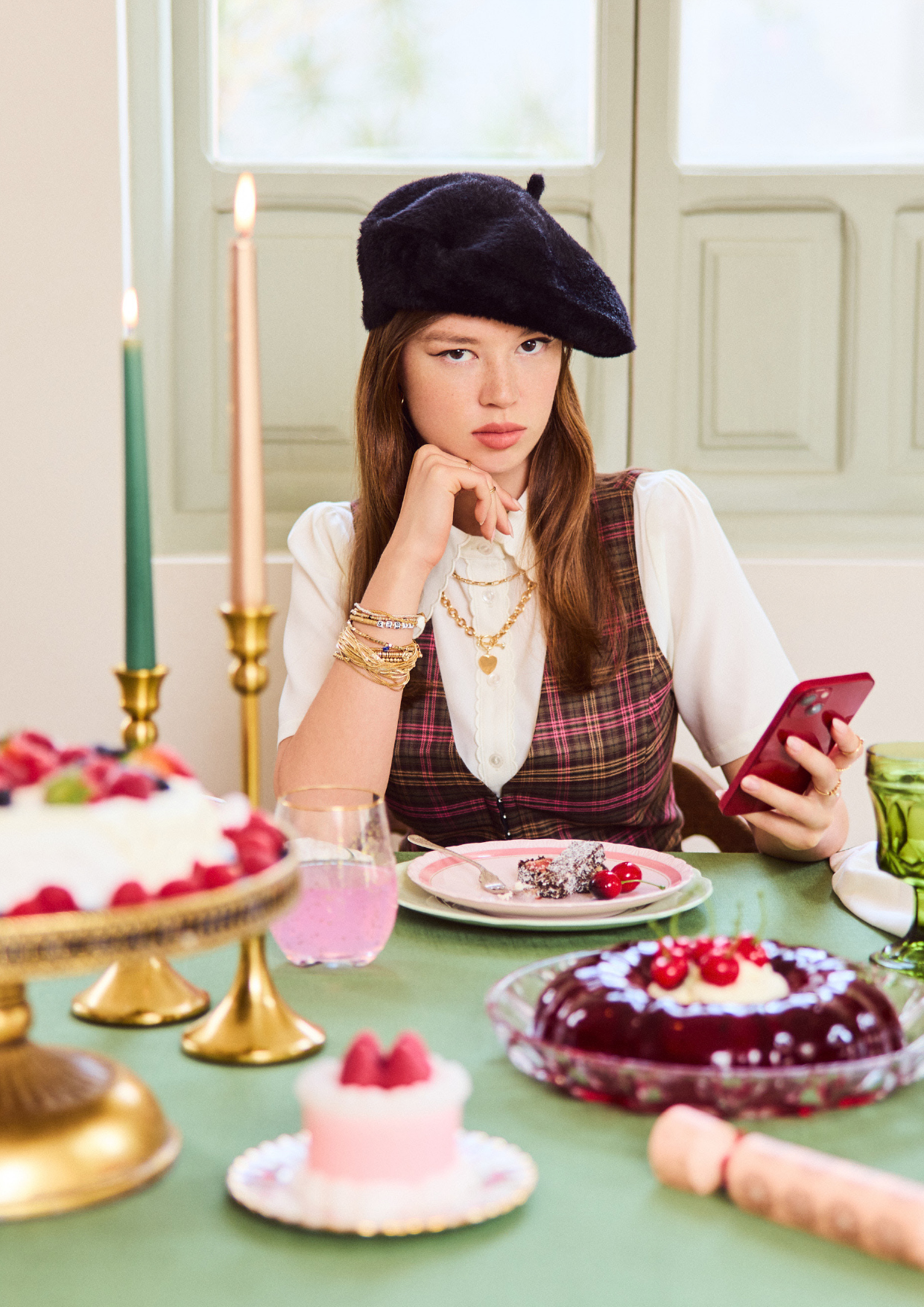 Young woman in beret sits at table with desserts, using red smartphone indoors with lit candles and green tablecloth. 
