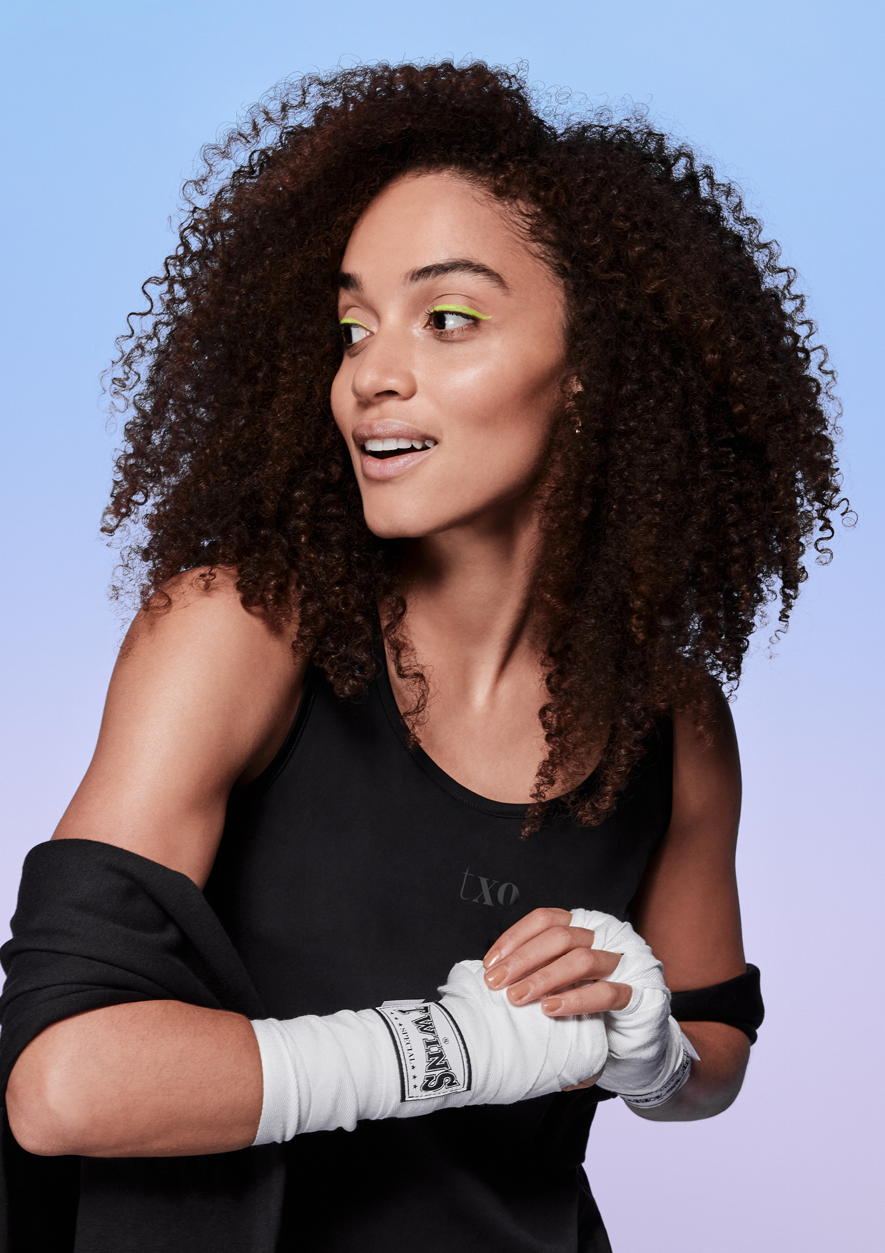 Young woman with curly brown hair wears boxing wraps and a black top against a gradient blue and pink background in a studio shot.