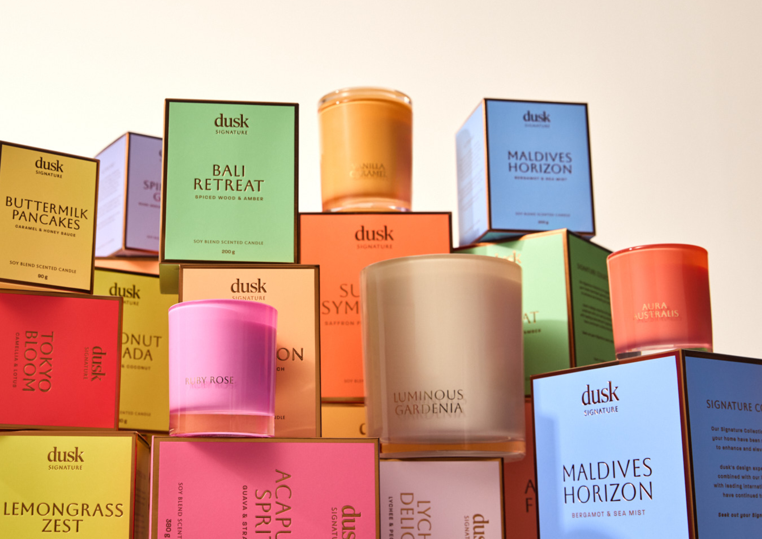Assorted 'dusk' brand scented candles and their packaging stacked in a studio environment with bright, soft lighting, product photography.