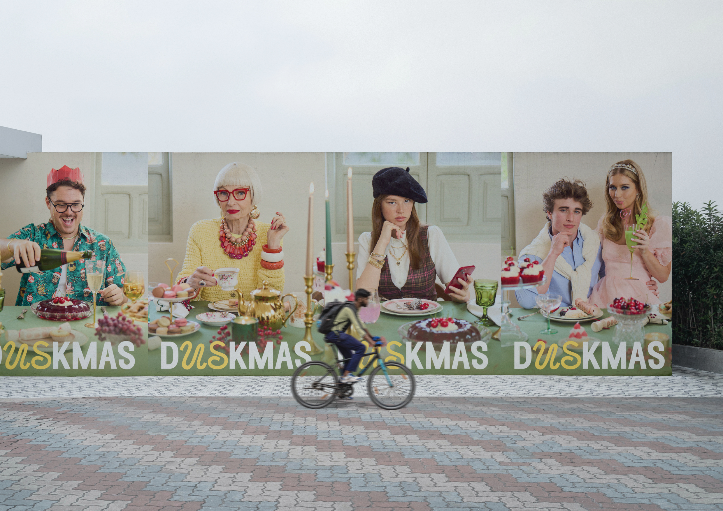 Outdoor billboard featuring four adults at a decorated table, with a cyclist passing by on a paved surface, sky visible above.