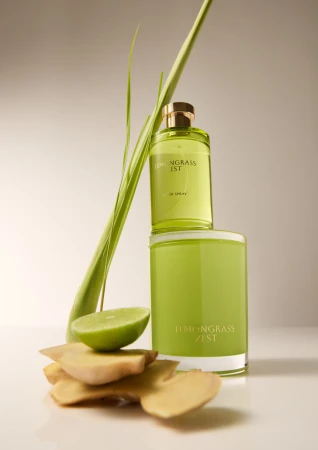 Still life shot of lemongrass-scented candle, room spray, lime, and ginger root against a neutral background in soft, diffused lighting.