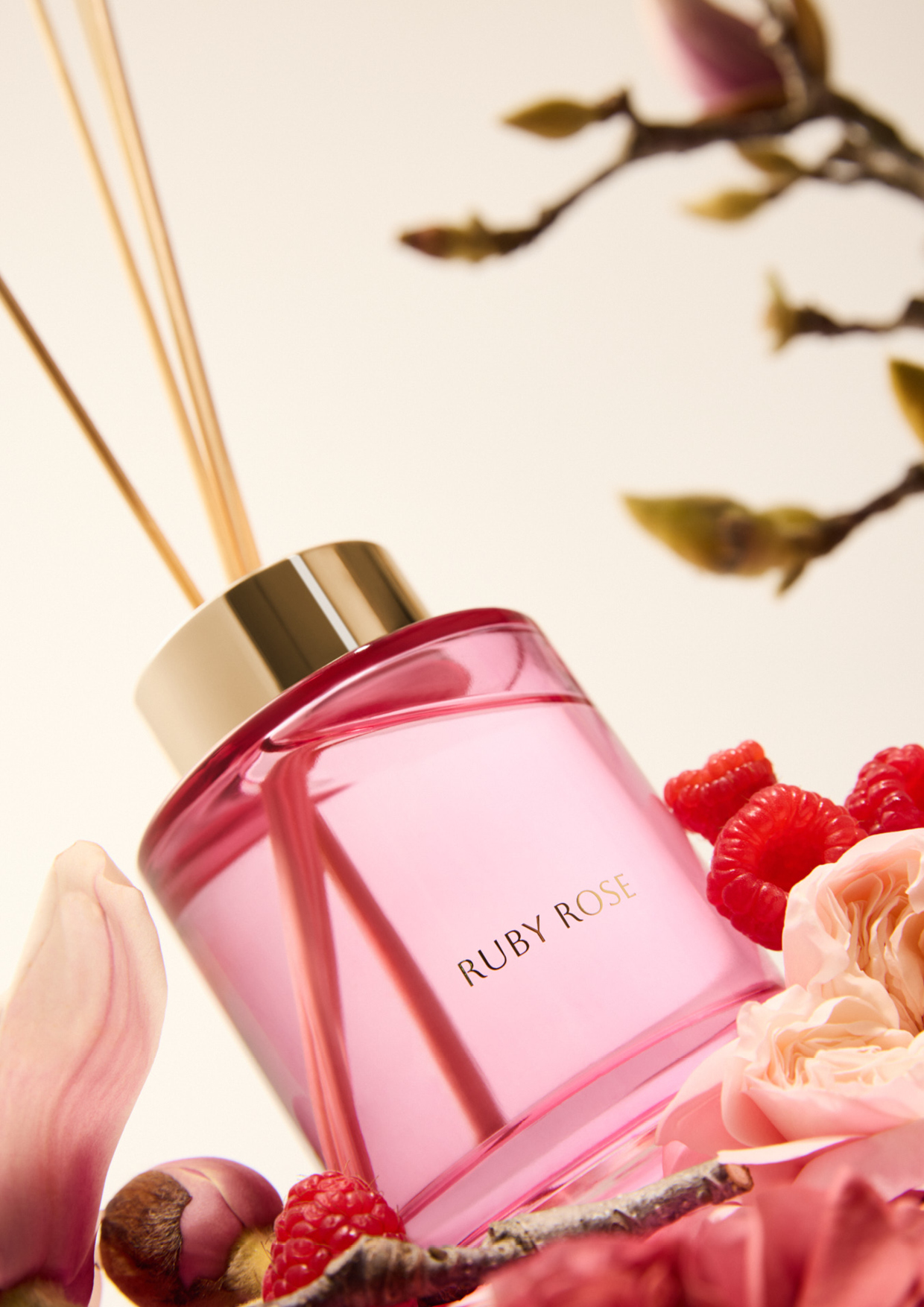 A luxurious room fragrance diffuser with a pink square bottle, gold top, and natural reeds is surrounded by magnolia blooms and raspberries.