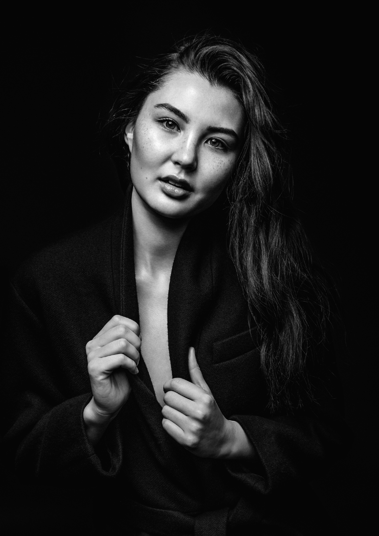 Monochrome portrait of a woman with dark hair wearing a dark blazer. Close-up shot against a black background with studio lighting.