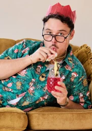 Man wearing a festive green shirt and red paper crown sipping a cocktail on a mustard sofa