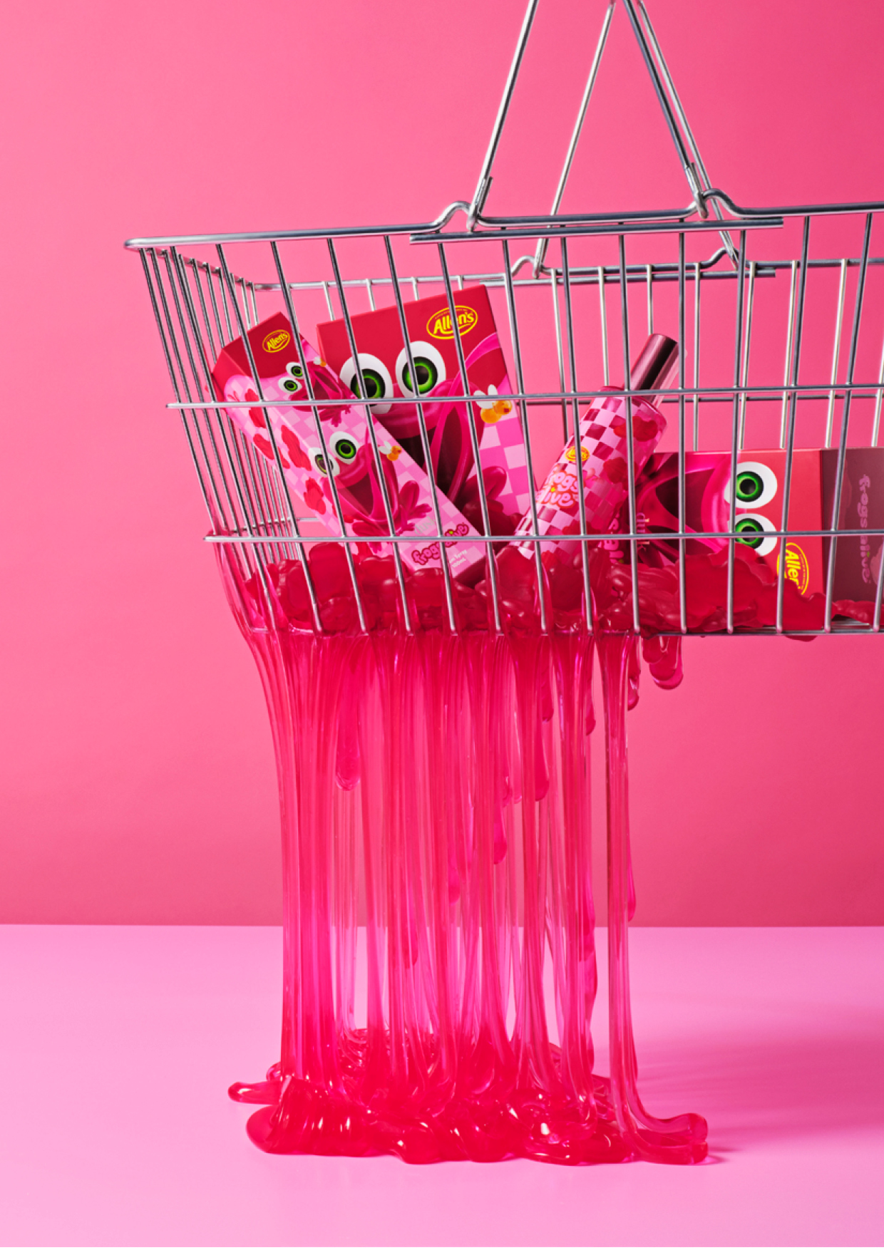 Silver wire shopping basket filled with pink Allens packaging and dripping vibrant pink slime against a matching pink background.