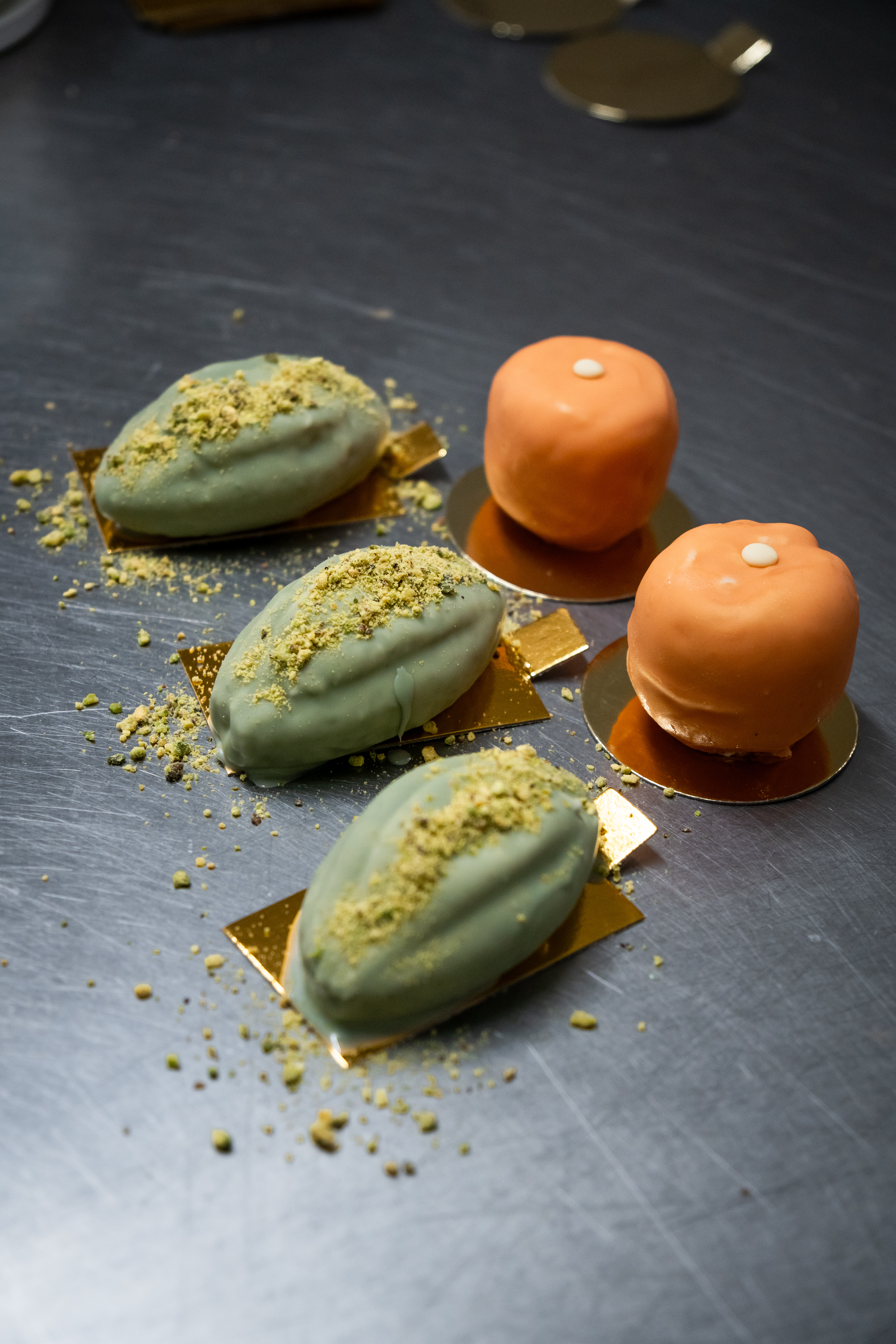 Green and orange petit fours on a gray surface. Food photography showcasing small, colourful pastries decorated with pistachio crumbs.
