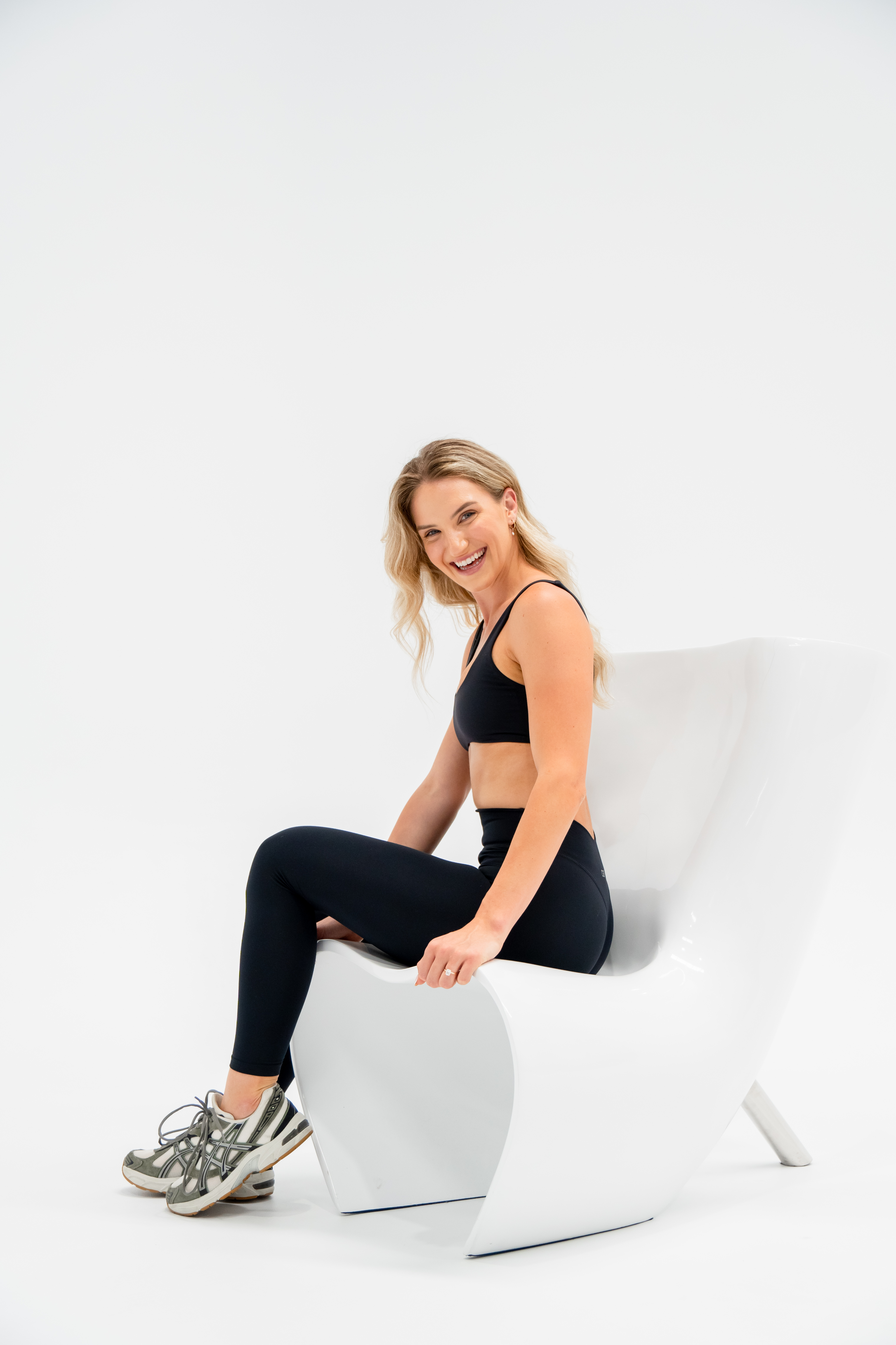 Smiling woman in athletic wear sitting on modern white chair in a white photo studio, looking at the camera in a portrait-style shot.