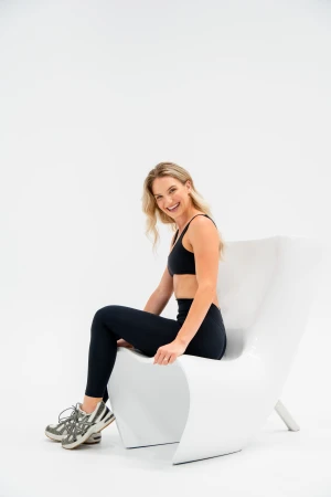 Smiling woman in athletic wear sitting on modern white chair in a white photo studio, looking at the camera in a portrait-style shot.
