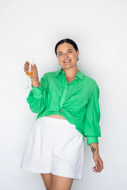 Smiling adult woman poses in green button-down shirt and white shorts, holding a champagne glass in a studio setting on white.