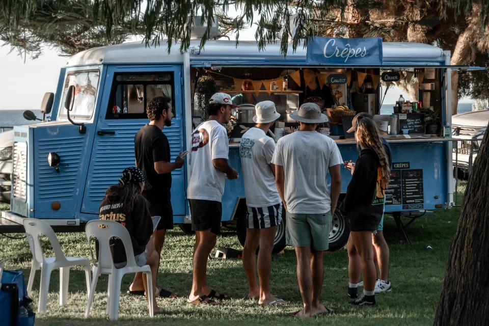 People gather at a light-blue food truck to purchase crêpes on a sunny day. White plastic chairs sit on the grass nearby.