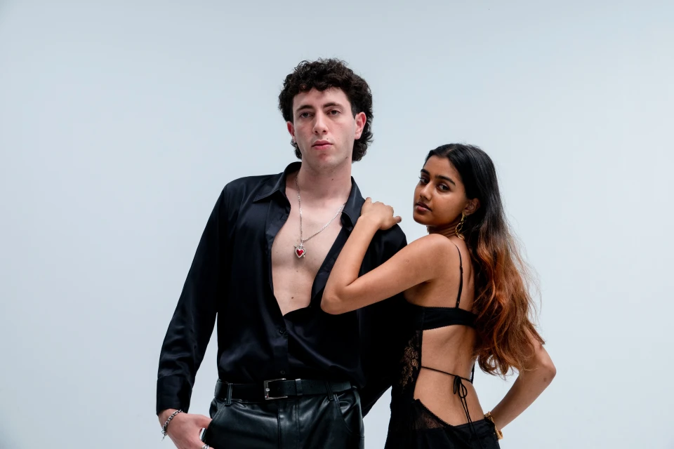 Attractively posed man and woman, wearing black clothing, set against a white studio background. Man is wearing a striking red pendant.