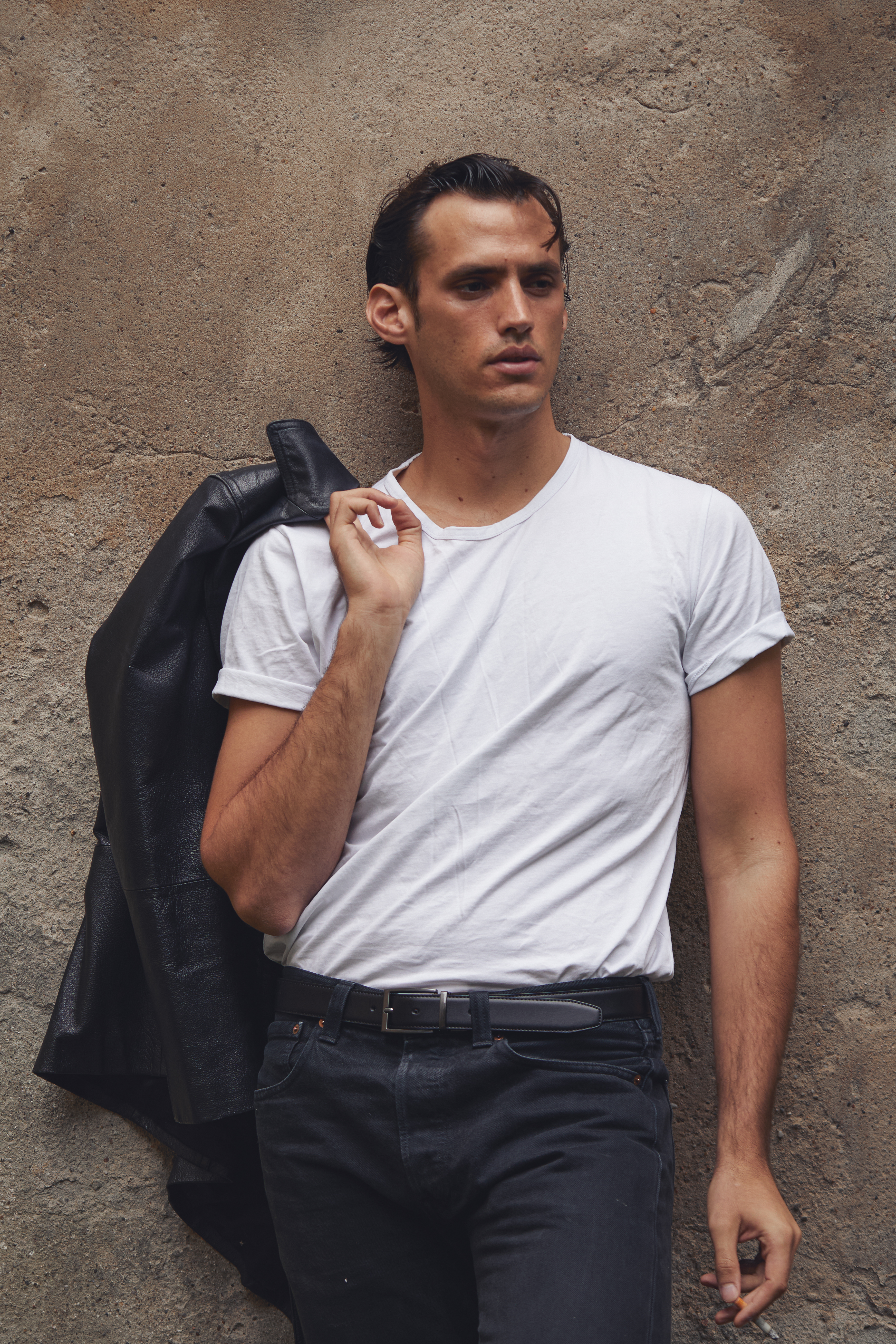 Man in a white t-shirt poses against a concrete wall with a leather jacket slung over his shoulder in an outdoor setting.