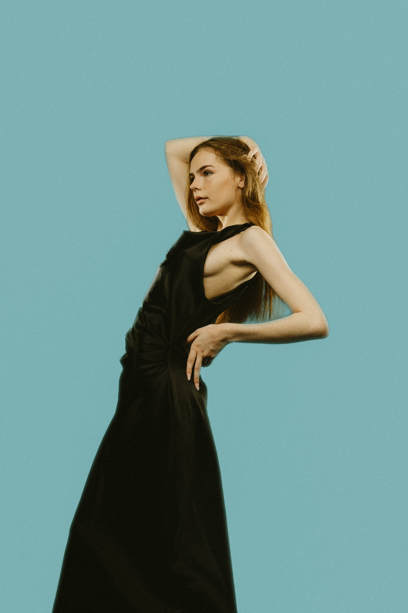 A woman in a long black dress poses against a light blue backdrop, her hand behind her head and her body arched back in a fashion pose.