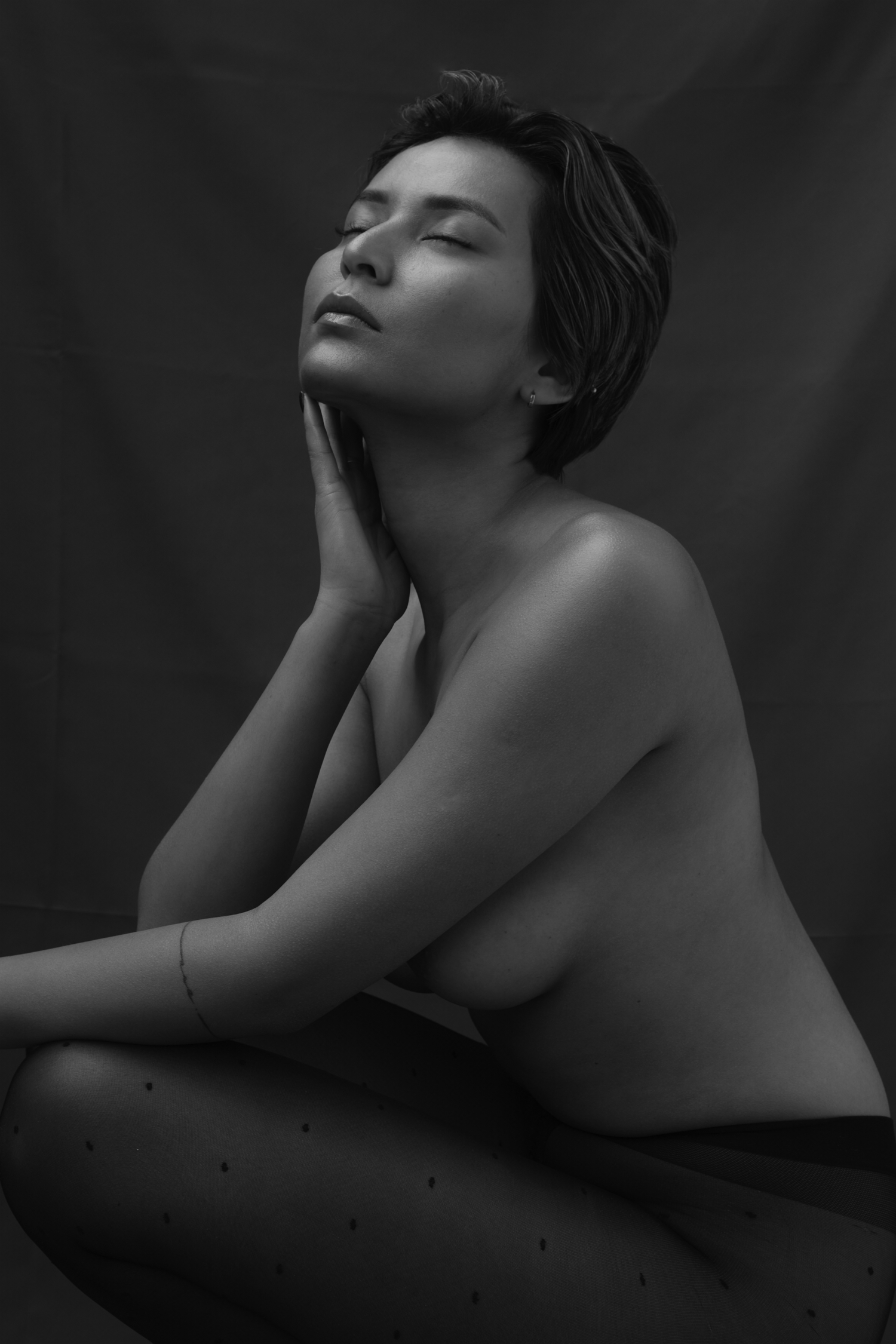 Monochrome portrait of an adult woman with short hair in dotted tights, eyes closed, and hand on her neck against a dark background.