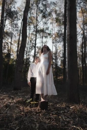 Man in white shirt and woman in white dress standing among tall trees in a forest, looking upward