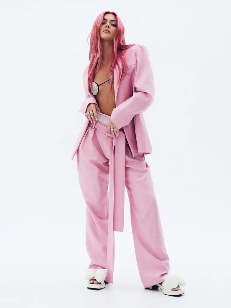 Full-body studio shot of a pink-haired adult female wearing a pink blazer and pants with white fluffy slide sandals against a white backdrop.