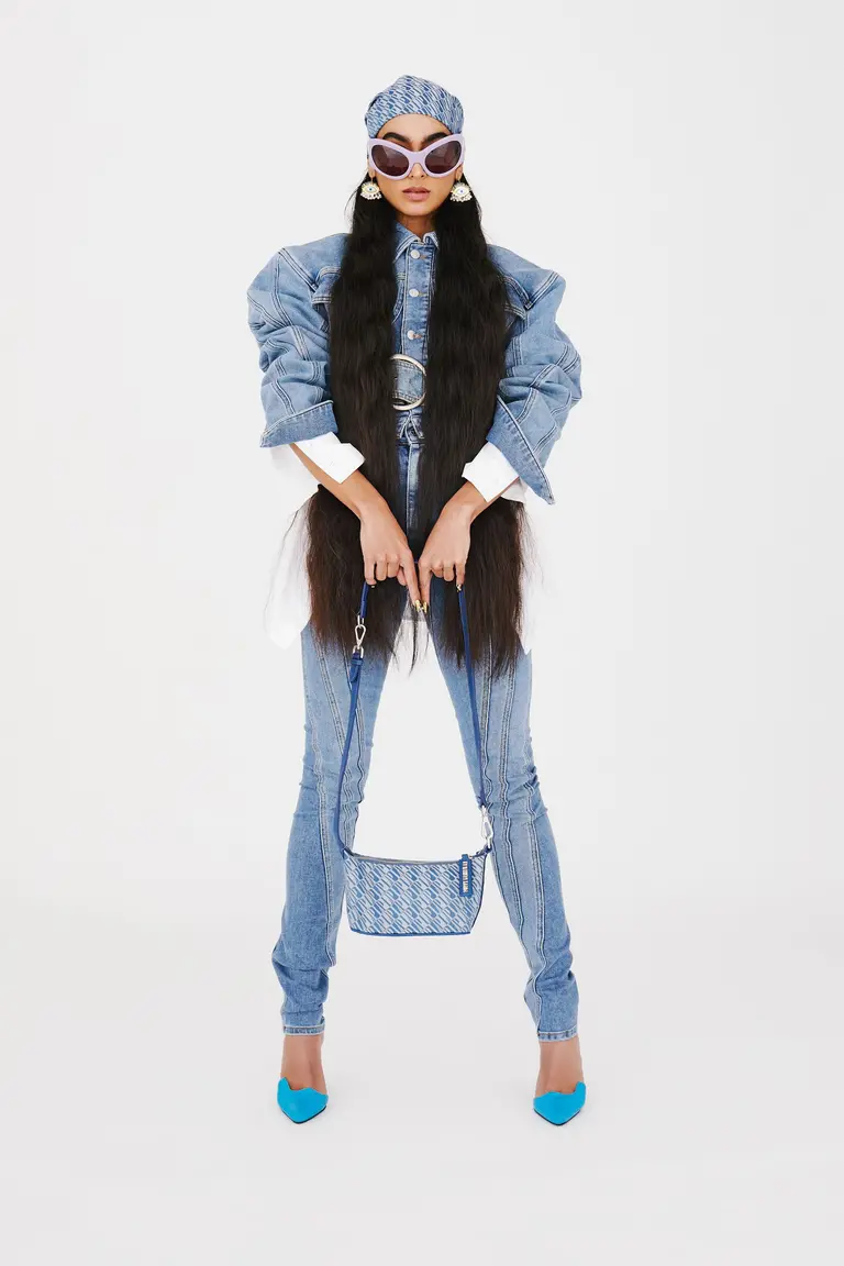 Fashionable woman in denim ensemble accessorized with sunglasses and handbag stands in a brightly lit, neutral-toned photo studio.