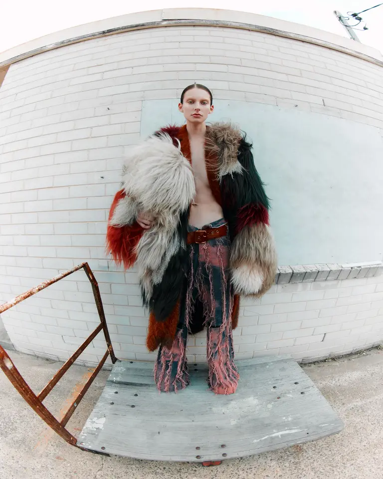 A person stands on a weathered platform, clad in patchwork fur coat and tattered pants, against a distressed white brick backdrop.