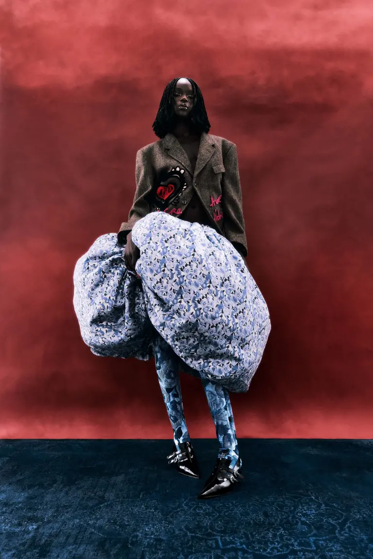 A Black female fashion model in a tan blazer and blue floral print dress poses in a studio against a textured red and blue backdrop.