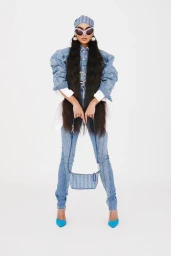Model in denim outfit wearing sunglasses and headscarf holding a shoulder bag against a white backdrop