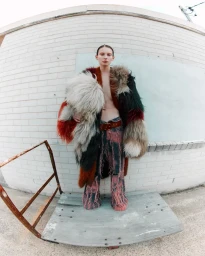 Model wearing a multicolored faux fur coat and fringed pants standing on a wooden platform against a white brick wall.