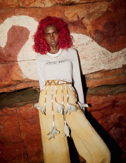 Fashion shot of a woman with red curly hair in front of a rock wall with cave paintings wearing a white long sleeve top and light brown pants.