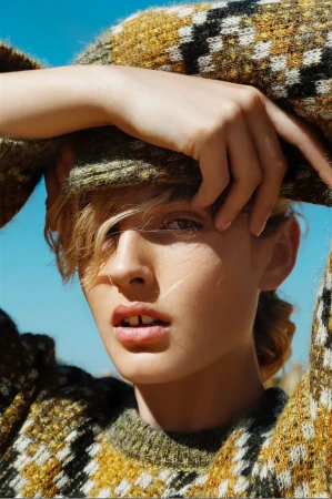 Close-up portrait of a woman wearing a patterned knit sweater with hands framing her face against a bright blue sky.