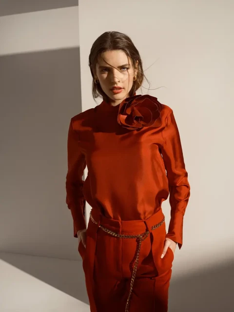 A brunette woman in a burnt orange silk blouse with a flower appliqué and matching pants stands in a photo studio with directional light.