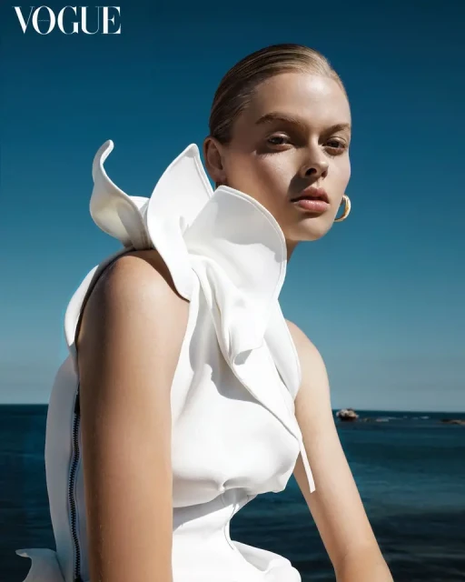 Young woman with slicked-back hair and hoop earrings poses in an architectural white dress with a high collar against a blue sky and sea.