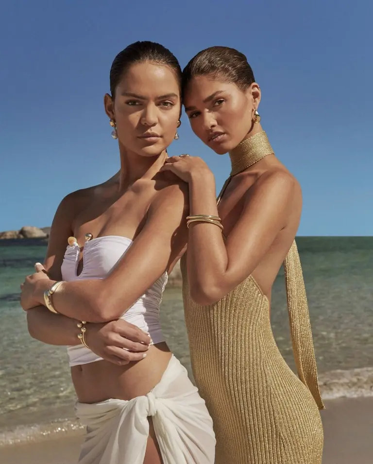 Two women in swimwear and jewelry pose on a sunny beach with clear sky and ocean backdrop, showcasing fashion and beach lifestyle.